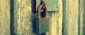 Close-up of a rusty padlock attached to a weathered wooden door, resembling those often found on old church entrances. The lock shows signs of wear and aging, enhancing its historical vibe. The softly blurred background highlights the padlock's texture and detail.