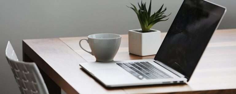 A laptop sits open on a wooden table next to a white coffee cup and a small potted plant, likely used to brainstorm ideas for church website improvements. A white chair is partially visible on the left, suggesting this tidy workspace is perfect for focusing on website tips.