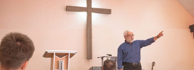 An older man with a beard, wearing a blue shirt, gestures while addressing a small group in a church setting. As the Executive Pastor, he stands confidently before them with a large wooden cross behind him. A podium and musical instruments are also visible in this serene atmosphere.