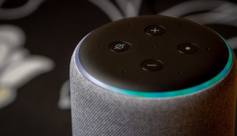 Close-up image of a smart speaker with a fabric exterior, reminiscent of Alexa's elegant design. The top features four control buttons, including volume up and down, and the device is lit with a blue ring indicator. A blurry floral pattern is in the dark background.