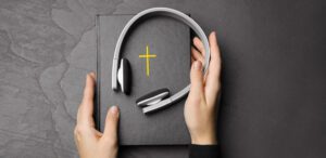 Hands hold a black book with a yellow cross on its cover, while gray and black headphones rest on top, ready to immerse you in streaming audio. The background is a textured gray surface.