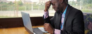A man in a suit sits at a wooden table with a laptop, resting his forehead on his hand while looking at the screen. He appears to be deep in thought about online church strategies to avoid burnout. A window with an outdoor view is in the background.