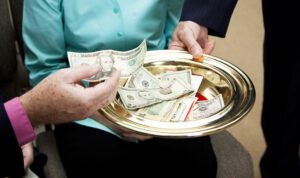 A person places a dollar bill into a gold-colored offering plate, contributing to church finances. The plate, containing various bills, is held by another person in a suit. They are seated indoors, likely within a religious or community setting.