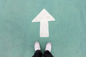 A pair of white sneakers stands on a green concrete surface, facing a large white arrow painted on the ground pointing forward—a vivid symbol of brilliance and the urge to go forth.