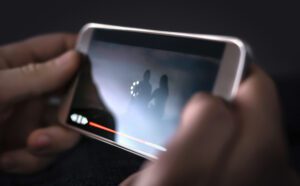A person holds a smartphone displaying a live streaming video with a buffering icon. The video timeline is partially red, indicating loading progress. The screen shows a blurred silhouette of two people.