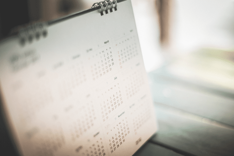 A close-up of a spiral-bound calendar open on a wooden table, highlighting the months of March and July. The scene captures productivity as it unfolds in soft, natural light, with blurred surroundings adding to the focus.