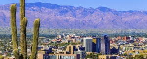 A vibrant cityscape of Tucson, Arizona, where modern buildings rise against rugged mountains. In the foreground, tall saguaro cacti reflect a thriving community fostering positive change under a clear blue sky.