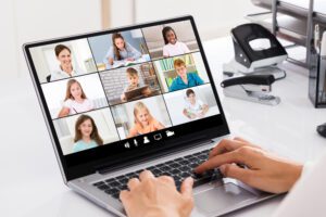 A person is typing on a laptop displaying a video call with nine participants, including adults and children, each in their own square. As a Great Church Host, they orchestrate the virtual gathering with grace. The background shows office supplies like a stapler and pen holder.