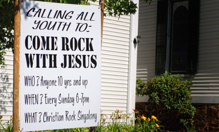 A sign outside a blooming church invites youth to "Come Rock With Jesus." Open to anyone 10 and up, the Christian Rock Singalong happens every Sunday from 6-7pm near a white-paneled building. A perfect example of effective church advertising.