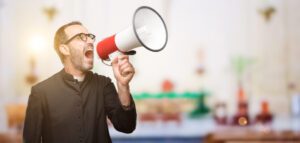 A man in a black clergy outfit is enthusiastically speaking into a red and white megaphone, aiming to improve church communication. The blurred background suggests a bright indoor setting, emphasizing the importance of clear church announcements.