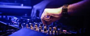 Close-up of a DJ's hand skillfully fixing audio on a sound mixer in a dimly lit, blue-toned setting, wearing a black wristwatch.