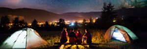 Four people sit on a bench between two tents at the church camp, gazing at a cityscape illuminated at night. The sky above is filled with stars, and distant hills form a serene backdrop.