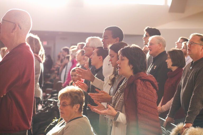 A diverse group of people, including the elderly, stands in a sunlit room engaged in an activity. Some have their hands raised and eyes closed, embodying the communal spirit akin to church attendance. The atmosphere is focused and unified, highlighting the benefits of shared experiences.