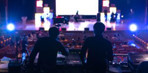 Two silhouetted church techs operate sound and lighting equipment at the back of a dimly lit auditorium. A stage is illuminated in the distance with musical instruments visible. Bright lights create a dramatic atmosphere, contrasting with moments that might make guests feel unwelcome.