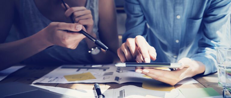 Two people are working together at a desk, crafting a strategy as they study the tablet screen. One points with a pen while the other uses their finger, surrounded by papers and a glass of water, setting the scene for innovation and improvement in 2021.