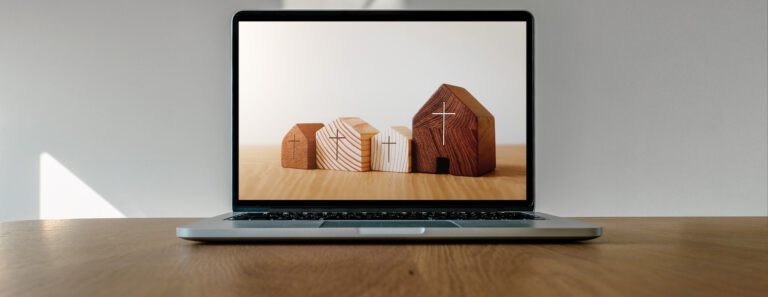A laptop rests on a wooden table, showcasing an image of four wooden blocks shaped like houses of varying sizes. The blocks have simple, etched details, reminiscent of a serene church website. Light streams in from the left, creating a peaceful ambiance against the soft beige background.