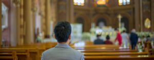 A person in a gray suit sits in a church pew, facing the altar. The ornate interior suggests it's welcoming to first-time visitors, with blurred figures in the background near the altar, possibly during a ceremony or service aimed at building visitor retention.