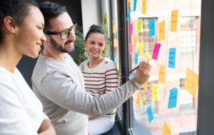 Three people are working together, smiling as they place colorful sticky notes on a window. The notes, filled with various handwritten ideas and words, suggest they're crafting a communication plan for their project in a bright room with large windows.