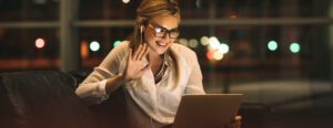A woman wearing glasses and earphones is sitting on a couch, smiling and waving at a laptop screen, virtually connected with members. The background shows blurred city lights through a window, suggesting it is nighttime.