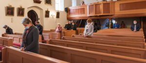 People wearing masks and practicing social distancing while standing and sitting in pews inside a church, as part of the Church Reopen Plan. Some are praying with heads bowed. The church interior shows wooden benches and framed pictures on the walls.