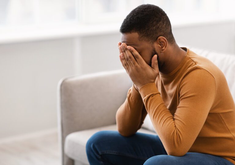 A person sitting on a couch covers their face with both hands, wearing a brown sweater and blue jeans. The bright window suggests it is daytime. The individual seems in deep contemplation, hinting at the pervasive issue of depression.