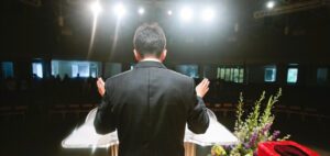 A person in a suit delivers tips from a podium adorned with flowers, their voice highlighted by bright lights in the dimly lit auditorium. A few unchurched guests linger faintly visible in the background.