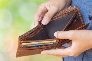 A person holds an open, empty brown leather wallet, hinting at church donations as several credit cards are visible in the card slots. The background is blurred with green and yellow hues.