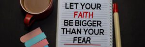 A notebook emblazoned with "Let your faith be bigger than your fear" sits ready for inspiration. Beside it, a red pen lies in wait, while a steaming mug of hope warms the left. Three colorful bookmarks offer encouragement and direction amid challenges like COVID-19.