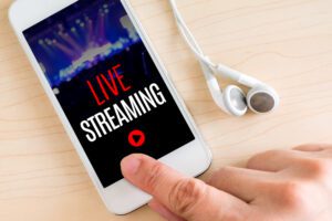 A smartphone showcasing a "Live Streaming" screen with a play button, ready to explore new ways to connect. The stage lights in the background blur beautifully, while a pair of white earphones lies nearby. A finger hovers, eager to dive into the stream.
