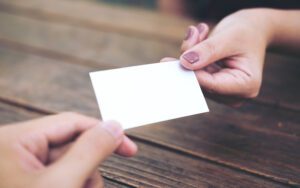 Two hands are shown exchanging a blank white church connection card over a wooden table. The card is held between the thumb and index fingers of each person, with natural light illuminating the scene, symbolizing a moment of meaningful church outreach.