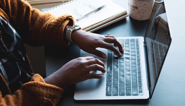 A person wearing a brown sweater is focused on blogging at a desk, typing on a laptop. A notebook, pen, and speckled coffee mug accompany their creative process, highlighting the benefits of mindful work.