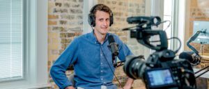 A person wearing headphones sits at a desk, sharing tips into a microphone. They are in a recording studio with brick walls, while a camera in the foreground captures them recording video content.