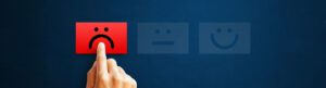 A hand is pressing a red button featuring a sad face emoji, symbolizing negative online reviews, while two unlit buttons depicting neutral and happy faces sit on a dark blue background.