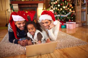 A family of three, donned in festive Santa hats and reindeer antlers, relaxes on a cozy rug in their living room. They're smiling at a laptop, enjoying a Christmas online service. In the background, a decorated tree and wrapped presents complete the joyful scene.