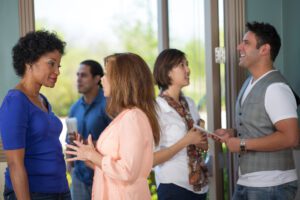 A small group of people socializing indoors exudes emotional intelligence. Two women in the foreground are engaged in thoughtful conversation, demonstrating their leadership qualities. In the background, a man and a woman chat and smile warmly as natural light pours through large windows.