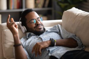 A man wearing glasses and headphones is relaxing on a couch with a content expression, gently raising one hand to the rhythm of music, enjoying live streaming audio. Shelves with books and plants are blurred in the background.