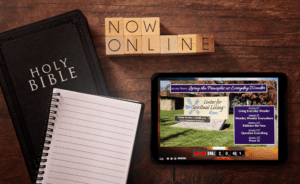 A Holy Bible and a spiral notebook sit on a wooden table beside a tablet streaming a church service from the "Center for Spiritual Living Reno" with the theme "Living the Principles as Everyday Decides." Scrabble tiles spell out "NOW ONLINE," inviting viewers to join in via video chat.