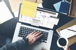 Sipping coffee, a person navigates their digital calendar planner on a laptop adorned with sticky notes and a small, paperclipped calendar. Amid notebooks and papers, they organize details for successful church events, ensuring every aspect of planning is seamless.