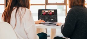 Two church tech experts sit at a table, collaborating on a video editing project using live encoders. The laptop screen shows editing software with various video clips. They're in a bright room with large windows casting natural light across their workspace.