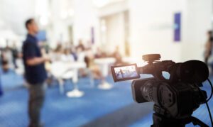 A video camera is filming a blurred scene in a bright, spacious room. A person stands out of focus in the background, among others, creating a lively atmosphere reminiscent of live streaming for churches. The camera's viewfinder captures intricate details of this vibrant setting.