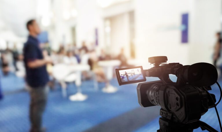 A video camera is filming a blurred scene in a bright, spacious room. A person stands out of focus in the background, among others, creating a lively atmosphere reminiscent of live streaming for churches. The camera's viewfinder captures intricate details of this vibrant setting.