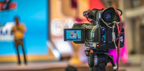 A close-up of a professional video camera on a tripod, capturing streaming tips from a blurred speaker on stage. The backdrop is vibrant, featuring shades of blue, orange, and pink.