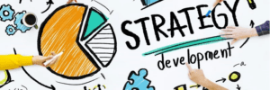 A collaborative work desk with hands pointing to a large "Strategy Development" text, highlighting Strategic Marketing. Surrounding are colorful doodles, including a pie chart, gears, and diagrams. Coffee cups and stationery add to the creative atmosphere.