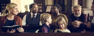 A diverse group of people sits in a church, engaged in conversation and reading. Two children in the front hold books, maximizing their understanding, while adults in the background, including a man in a suit and a woman with a scarf, reflect on the impact of the sermon as they interact warmly.