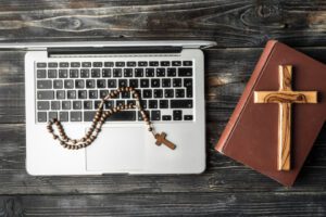 A rosary with a wooden cross rests on a laptop keyboard, ready for live streaming, next to a closed book with another wooden cross on its cover. This scene is set on a dark wooden table, symbolizing the ministry dedicated to bridging tradition with modern technology.