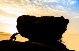 A silhouette of a person pushing against a large boulder on a hill at sunrise symbolizes the determination often seen in church development. The sky is a gradient of warm orange and cool blue, with scattered clouds representing the hindrances faced in achieving church growth.