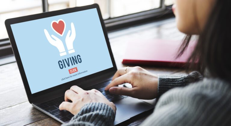 A person sits at a desk, typing on a laptop. The screen displays an image of two hands holding a red heart with "GIVING" below it and a clickable button that reads "click here to see online fundraising steps.