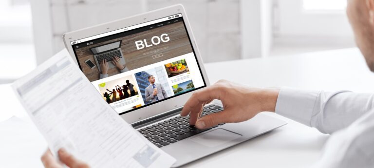 A person sitting at a desk looks at a laptop displaying a church blog with images and text. They hold a paper document in their left hand while utilizing the laptop touchpad with their right hand. The setting appears to be an office or study space.