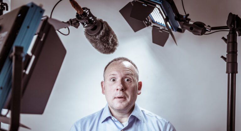 A person in a light blue shirt looks surprised, facing the camera. A large microphone is positioned above their head amidst professional studio lights, creating an atmosphere where even pastors could feel relaxed. The background is plain and softly lit.