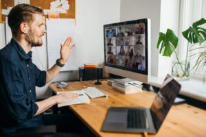 A man with earphones gestures while discussing the Pastor's Checklist in a video conference call on a desktop computer. He sits at a wooden desk with a notebook, pens, and a laptop. A potted plant and window provide a serene backdrop as he delves into church planning for 2021.